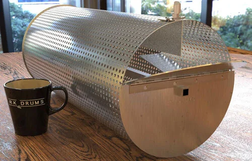 An RK Drums roaster drum on the table beside a branded RK Drums mug